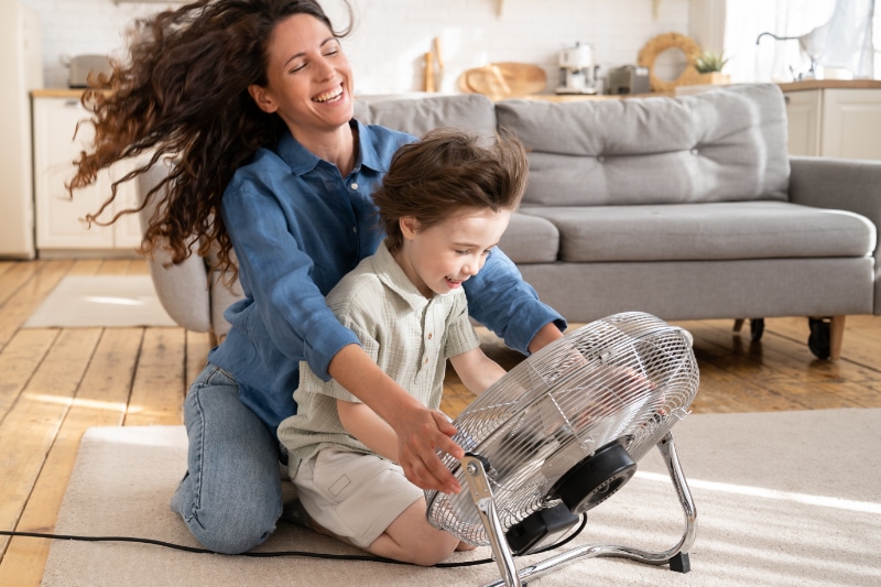 Loving mom relax and having fun with child at home: carefree young woman mother sit laughing together with son in front of fan ventilator work indoors and blow cool air during summer heat outdoors.