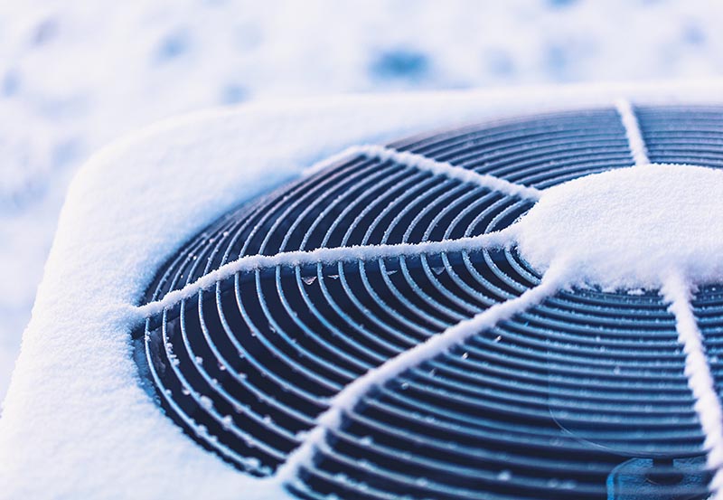 A close-up, top-down view of an outdoor air conditioning unit covered in fresh white snow and frost, with snow clinging to its circular metal fan grille.