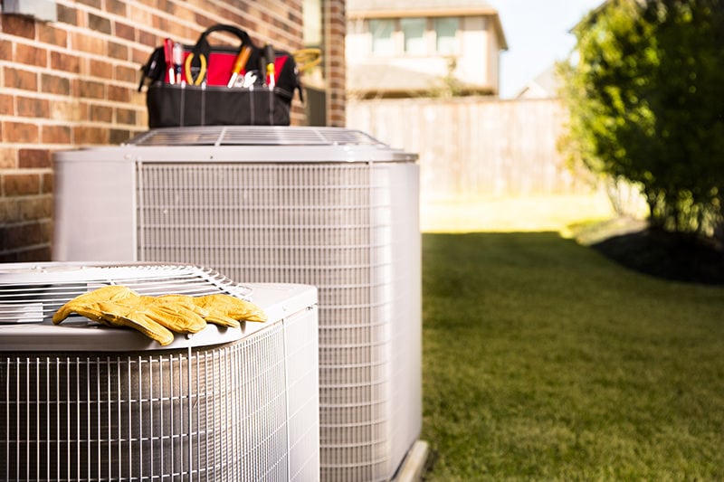 Benefits of Coil Cleaning and HVAC Maintenance. Bag of repairman's work tools, gloves on top of air conditioner units outside a brick home.