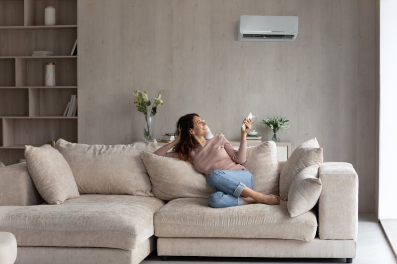 Planning to Remodel? Go Ductless! Relaxed young female homeowner sitting on huge comfortable couch, turning on air conditioner with remote controller, switching on cooler system, setting comfortable temperature in living room.