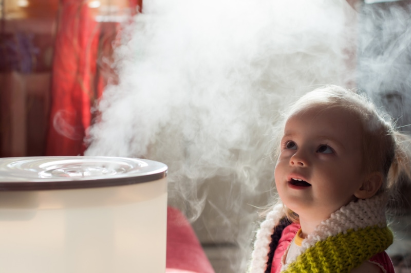 Benefits of Using a Humidifier in Your Home. Image is a photograph of a young girl looking in awe at an active humidifier.