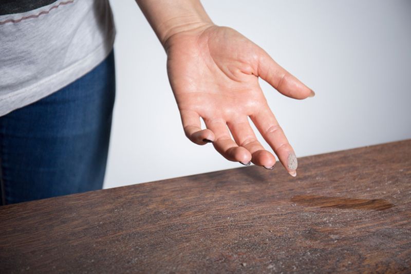 How to Reduce Dust in Your Home. Image is a photograph of a person's hand face-up over a dusty table. There is dust on the pad of their pointer finger.